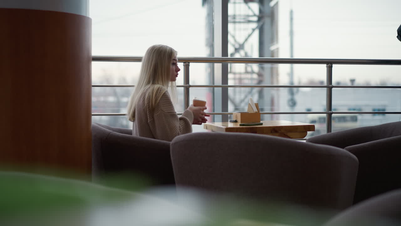 vista lateral de una joven con una taza de café, bebiendo una bebida en un acogedor café del centro comercial con el paisaje urbano borroso a través de la ventana, disfrutando de un momento tranquilo con la atmósfera urbana