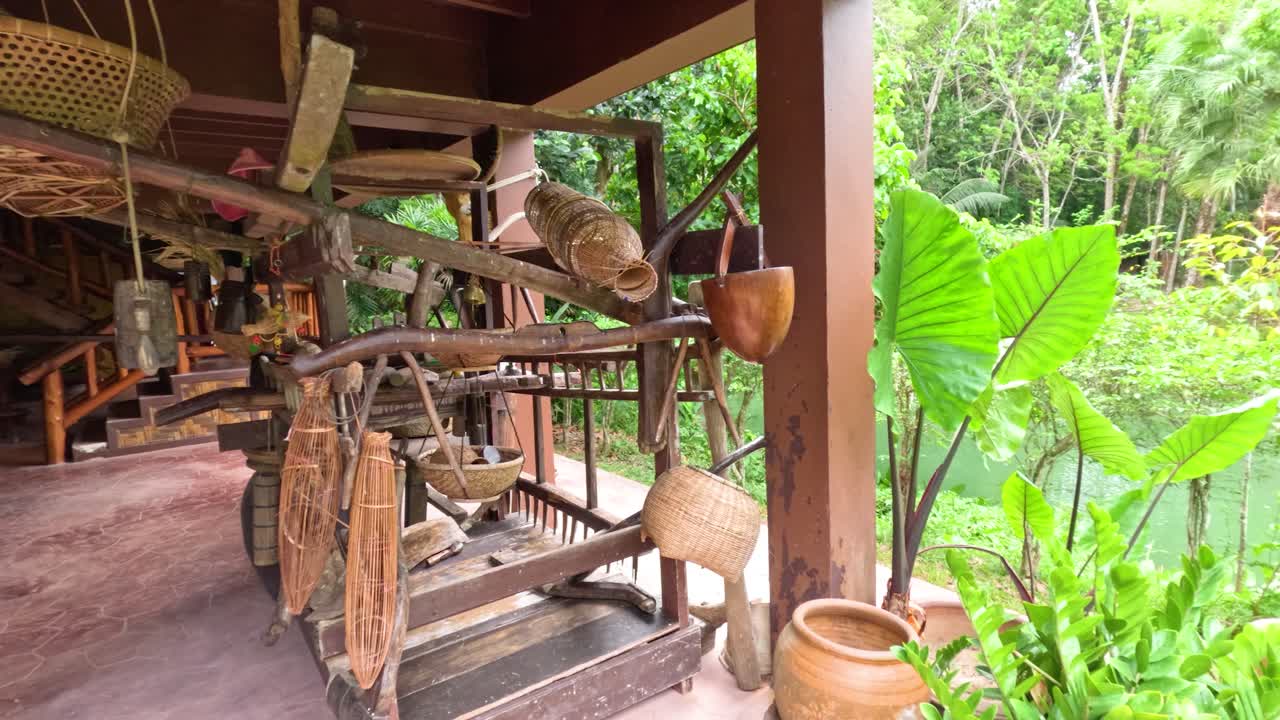Camera slowly pans past traditional Thai farming tools and baskets in open-air wooden house