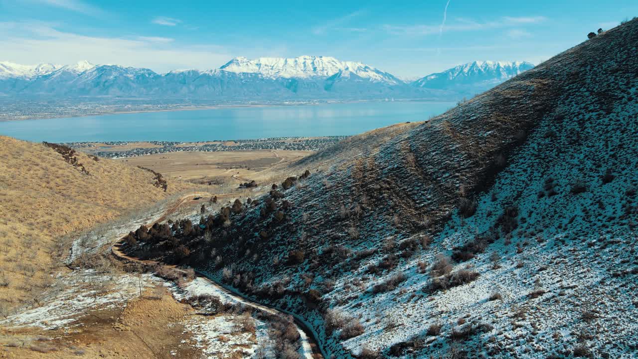 un camino de tierra a través de un cañón que conduce a una ciudad en el valle con montañas nevadas a través de un lago - vista aérea de retirada