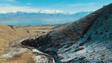A-dirt-road-through-a-canyon-leading-to-a-town-in-the-valley-with-snow-capped-mountains-across-a-lake---aerial-pullback-view
