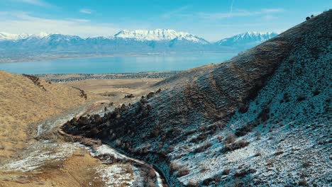 a dirt road through a canyon leading to a town in the valley with snow-capped mountains across a lake - aerial pullback view