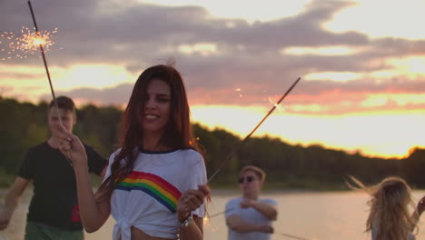 The-women-with-nude-waist-and-short-t-shirt-is-dancing-with-big-bengal-lights-in-her-hands-on-the-sand-beach-with-her-friends.-This-is-theamy-summer-evening-on-the-open-air-party.