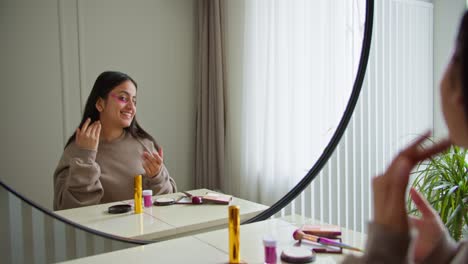 una chica morena con un suéter marrón aplica parches cosméticos especiales en su cara debajo de sus ojos para hidratar la piel y darle un color natural mientras se maquilla y mira su piel en un apartamento moderno durante el día