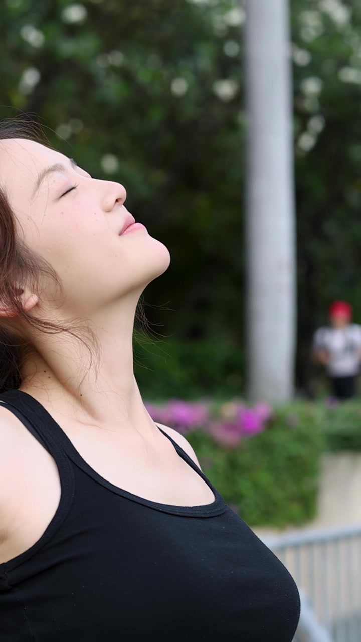 Uma mulher de roupa esportiva preta alonga o pescoço com os olhos fechados em um parque exuberante.