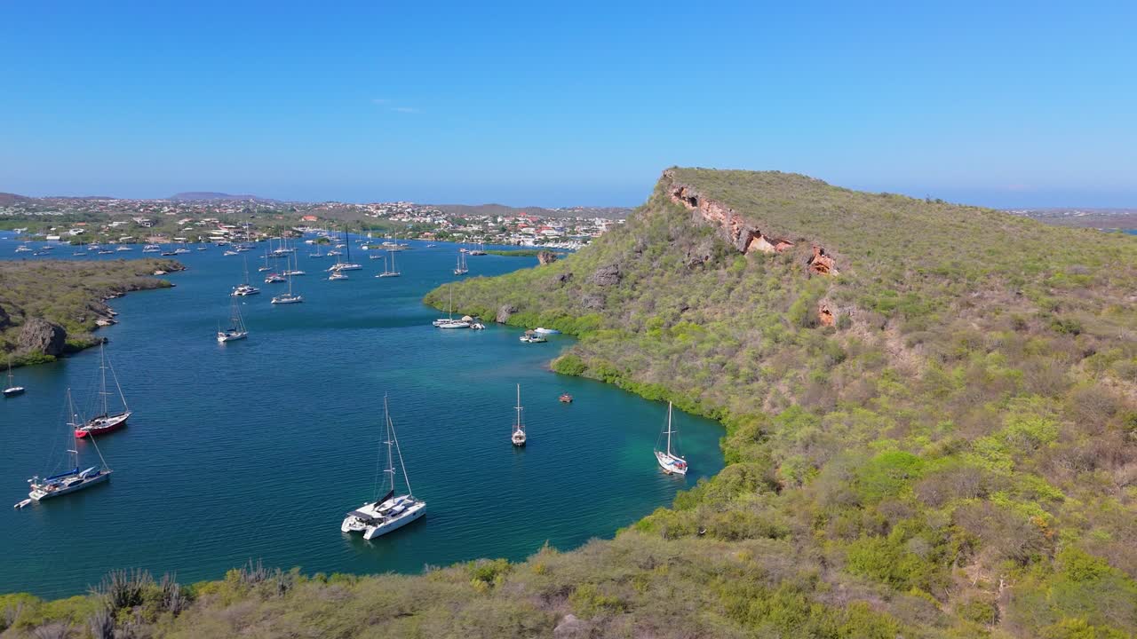 High-angle aerial of yachts anchored in clear waters near a rocky coast, with a distant hilly horizon in view