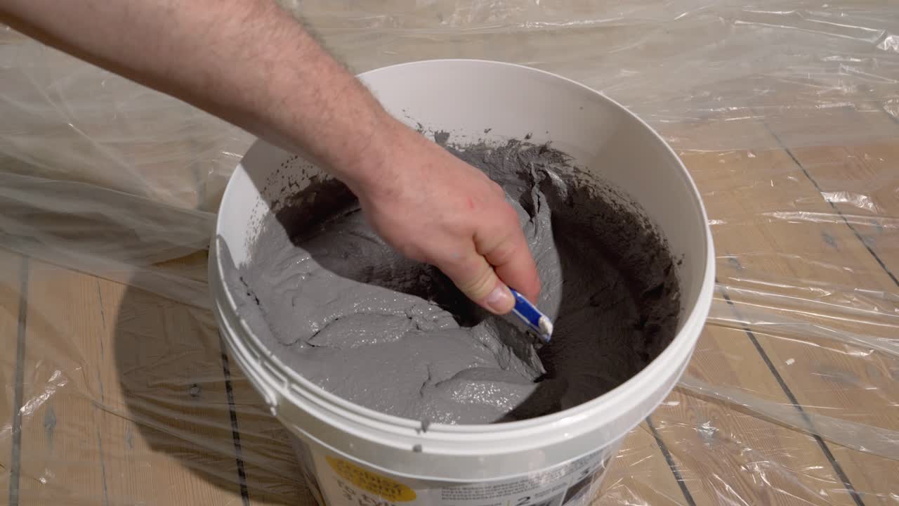 A bucket of decorative plaster stands on a wooden floor covered with painter's film, a man is mixing the plaster