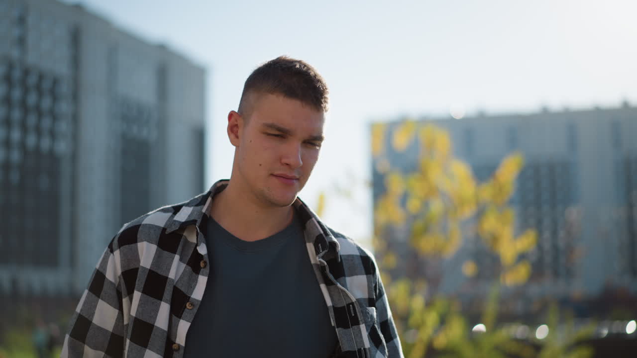 White young man in checkered shirt walks outdoors during sunny day with hands in pockets looking down thoughtfully in urban setting with blurred tall building and bokeh light in background