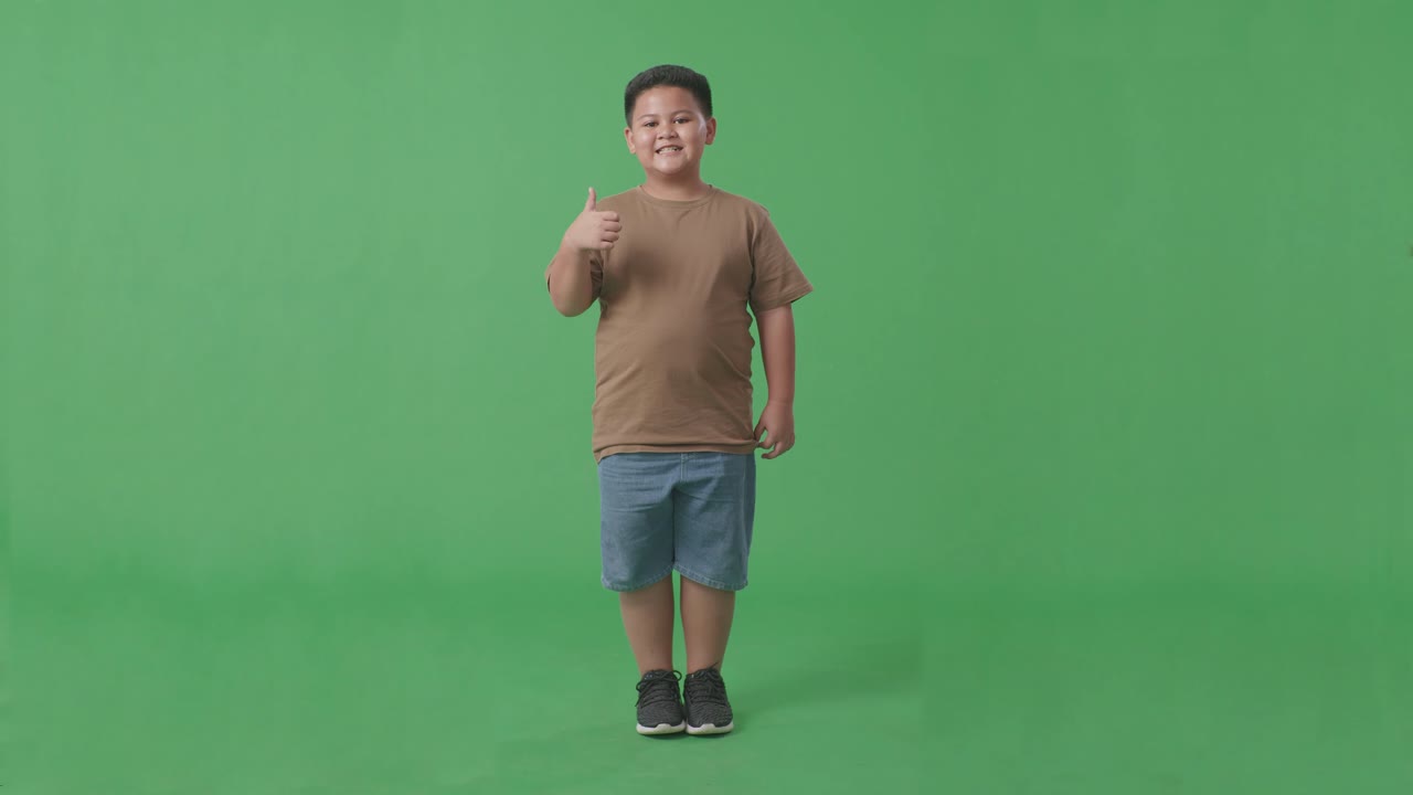 녹색 스크린 스튜디오에 서 있는 동안 엄지손가락을 올려 놓고 미소 짓는 아시아 소년의 몸이 가득 차 있습니다.