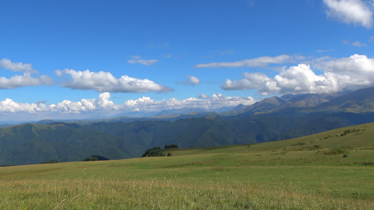ロシア、コーカサス山脈のドンバイにある、緑豊かな高山植物の牧草地と重なり合う山々の尾根が、明るい青空と雲の下に広がるパノラマビュー。