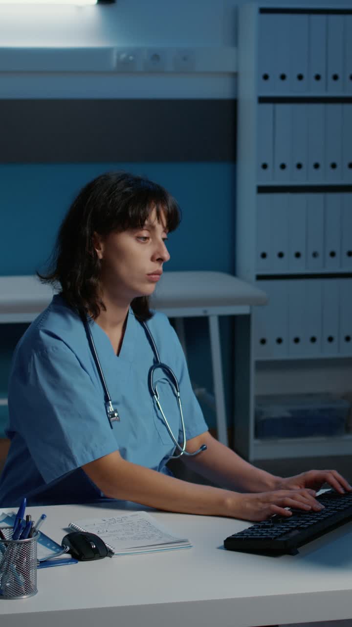 Vertical video: Exhausted nurse in blue uniform working over hours in hospital office