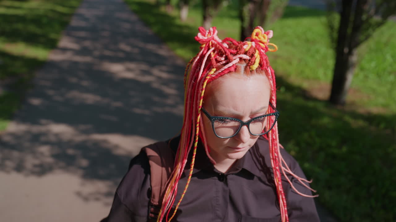 Estudiante reflexiva de piel blanca paseando por un parque, mirando su smartphone, con trenzas rojas y amarillas recogidas, camiseta negra y correas de mochila, luz solar moteada, tranquila vegetación urbana, expresión reflexiva