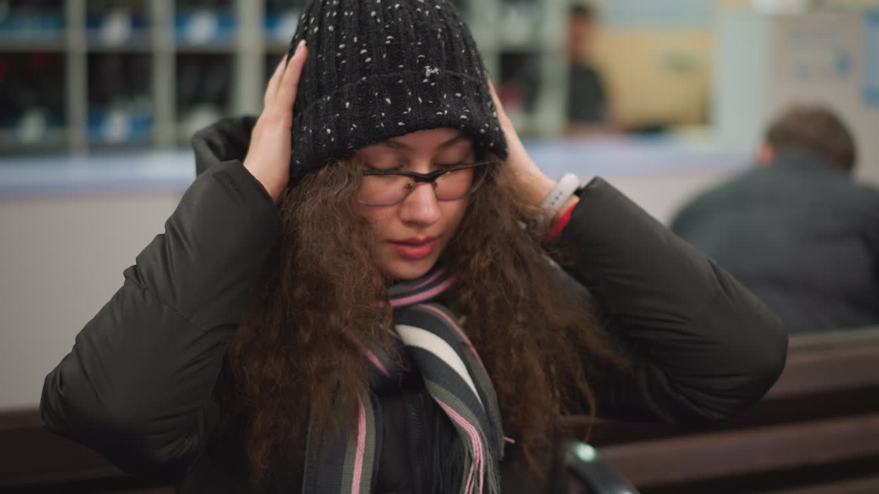 Damsel in glasses with long curly hair wearing black winter jacket and striped scarf looks downward and wears her head warmer clothing indoors, everyday lifestyle expression