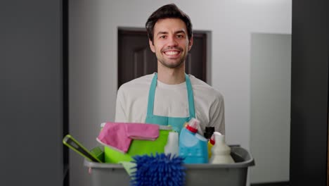 Porträt-Eines-Selbstbewussten-Brünetten-Hausmeisters-In-Blauer-Uniform-Und-Weißem-T-Shirt,-Der-In-Seinen-Händen-Ein-Graues-Und-Plastisches-Becken-Hält,-Das-Mit-Vielen-Werkzeugen-Zum-Reinigen-Der-Wohnung-In-Einer-Modernen-Wohnung-Gefüllt-Ist