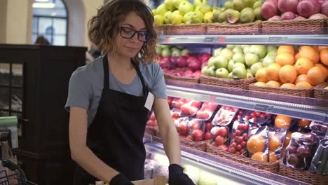 close up of pretty caucasian worker in black apron and gloves stocking the fruits in supermarket. young employee in glasses at