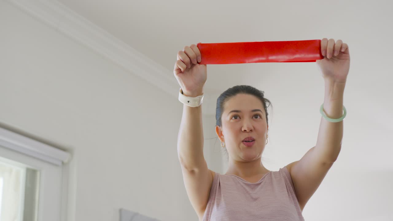 Donna asiatica che si allena e tira una fascia di resistenza rossa sopra la testa a casa, indossando uno smartwatch bianco.