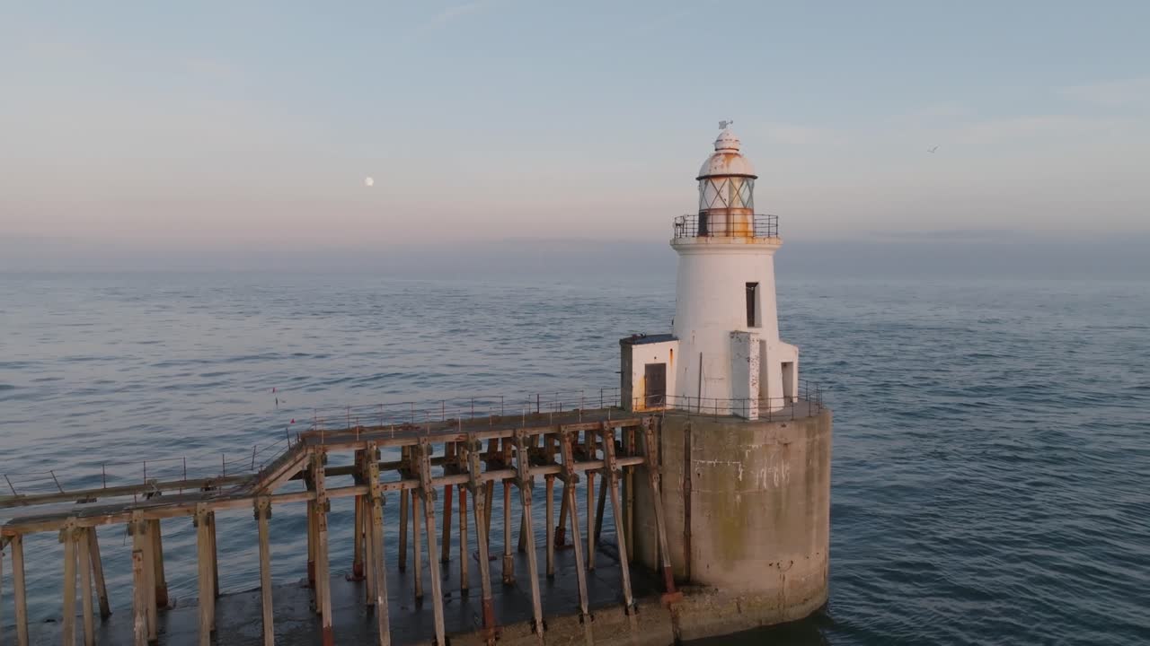 White lighthouse with rustic jetty at golden hour sunset, surrounded by calm seas and pastel skies