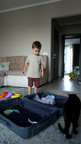 little baby boy packs a big grey suitcase lies opened on the floor. cute kid throws his clothes into the suitcase and closes it. black cat walks around. vertical video.