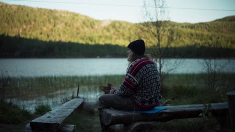 man sitting on tree trunk bench while drinking coffee by the lake in the morning