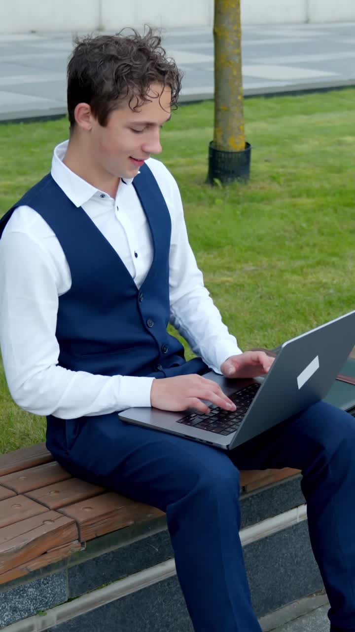 Young man in suit using laptop outdoors, looking focused and relaxed, vertical