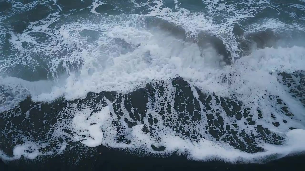 Breaking surf zone pushing incoming swell onto dark wet sand, forming cellular foam patterns