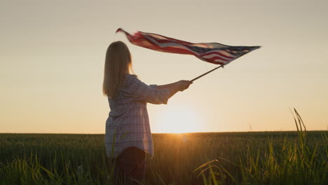 una mujer joven agitando la bandera de los estados unidos, de pie en el fondo de un pintoresco campo de trigo donde se pone el sol. video en cámara lenta