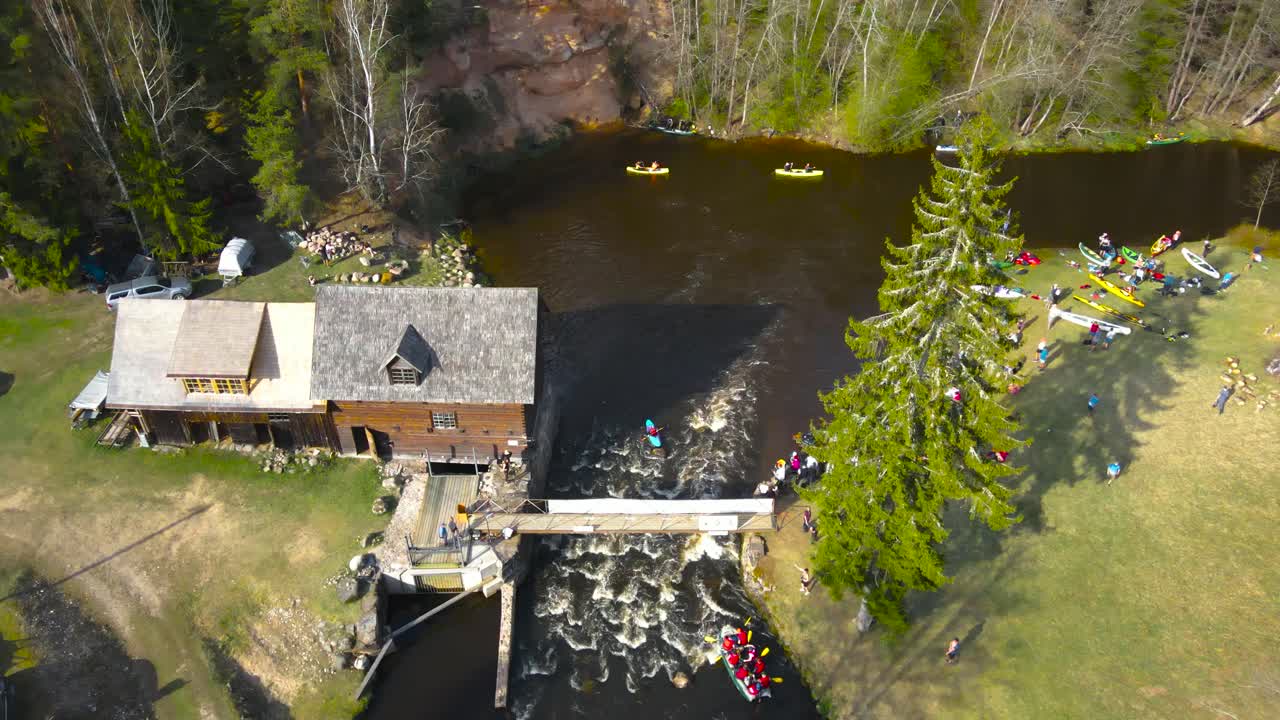 Aerial drone footage following and flying behind a large SUP paddleboard or a raft that is going through foamy narrow river rapids from underneath a bridge during a sunny day at Võhandu marathon.