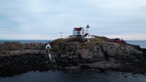 lighthouse on a rocky island at dusk with soft clouds in the sky and red rooves on buildings, tides crashing on the coast