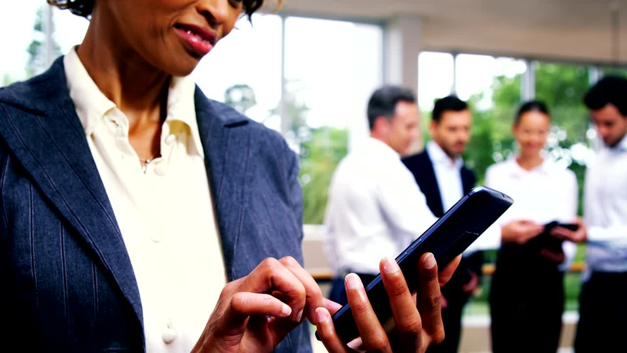 ejecutiva de negocios enviando mensajes de texto en el teléfono móvil mientras sus colegas interactúan en segundo plano