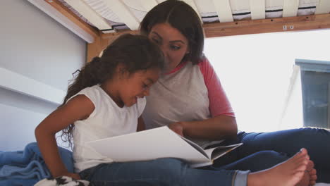 hispanic mother and daughter sitting on girls bunk bed at home reading book together