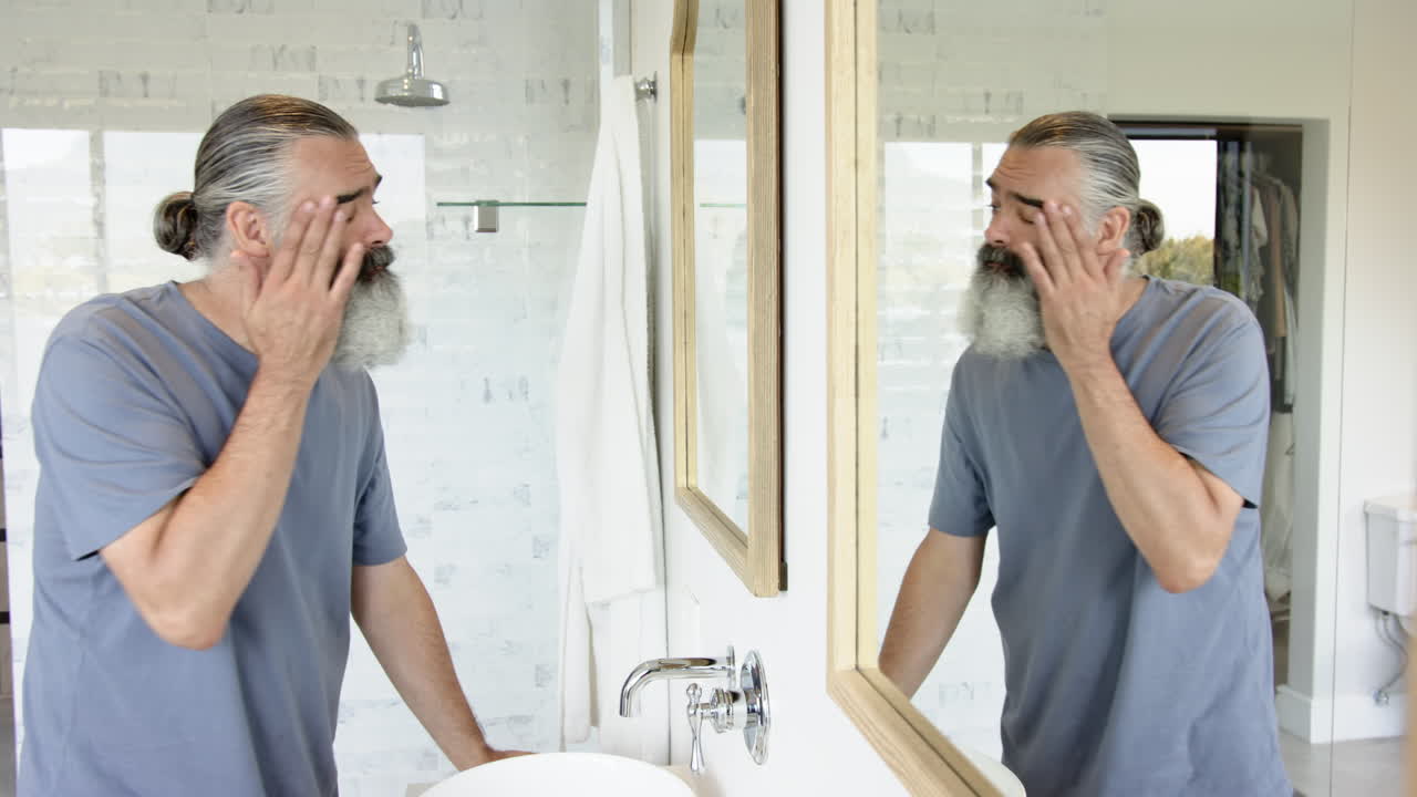 Man with gray beard looking at reflection in bathroom mirror, appearing thoughtful
