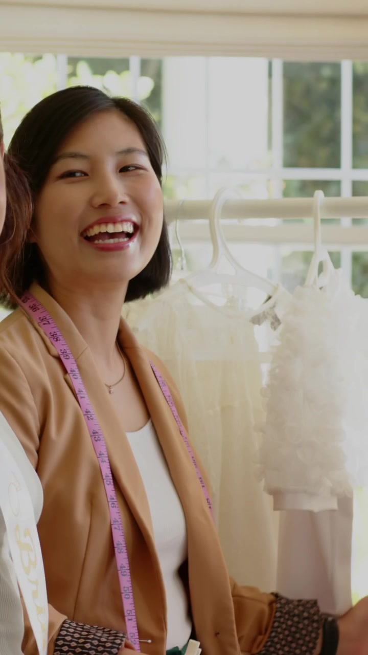 縦型動画：店で抱擁の後、クライアントがガウンを選ぶのを手伝うアジア人女性スタイリスト（ブレザー、パープルのテープ）