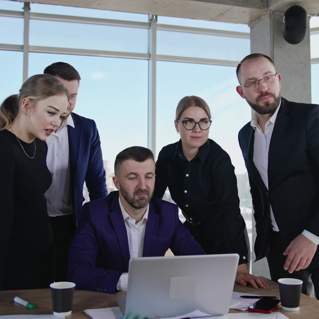 Office team standing around desk with laptop. Another man joins them after phone conversation. Sunny cityscape at the backdrop