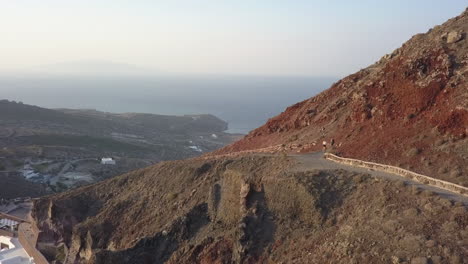 Aerial-tracks-two-hikers-walking-on-mountain-path-on-Santorini,-Greece