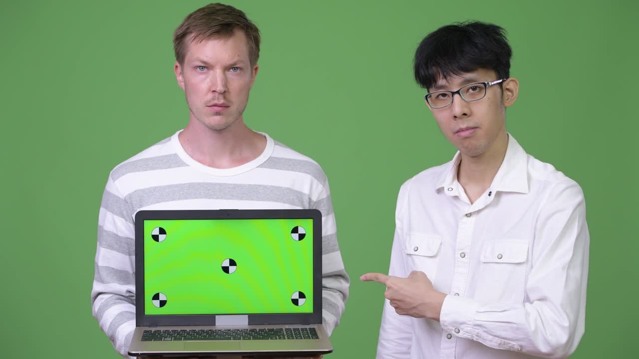 두 명의 젊은 다민족 사업가들이 손가락을 가리키면서 카메라에 노트북을 보여주고 있습니다.