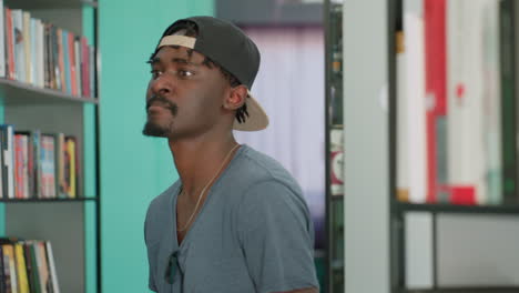Student in cap walks between library shelves, glancing at books with focused expression, teal wall background, calm academic atmosphere, casual attire, relaxed body language, exploring titles