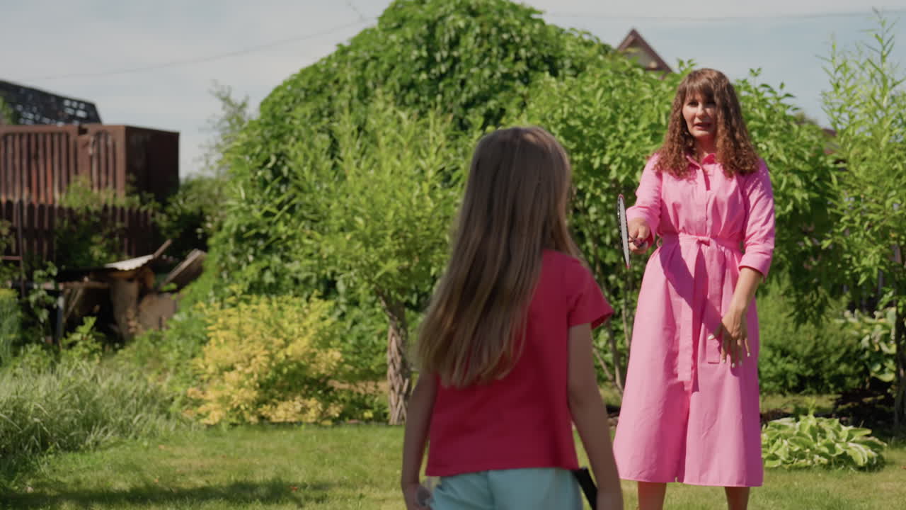 Caucasian Mother And Daughter Greeting In Sunlit Garden, Mother In Pink Dress Welcomes Young Girl Holding Racket, Lush Hedges And Lawn, Tender Hug And Smiling Faces, Joyful Family Moment