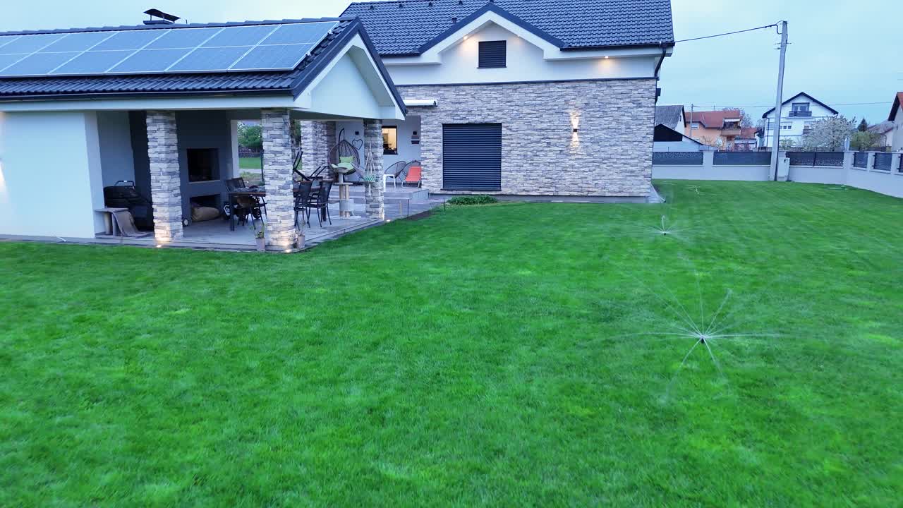 Zoom out udara dari rumah pintar modern saat senja. Rekaman drone memperlihatkan panel surya, arsitektur batu, dan penyiram rumput otomatis menyirami taman hijau di kawasan perumahan mewah pinggir kota