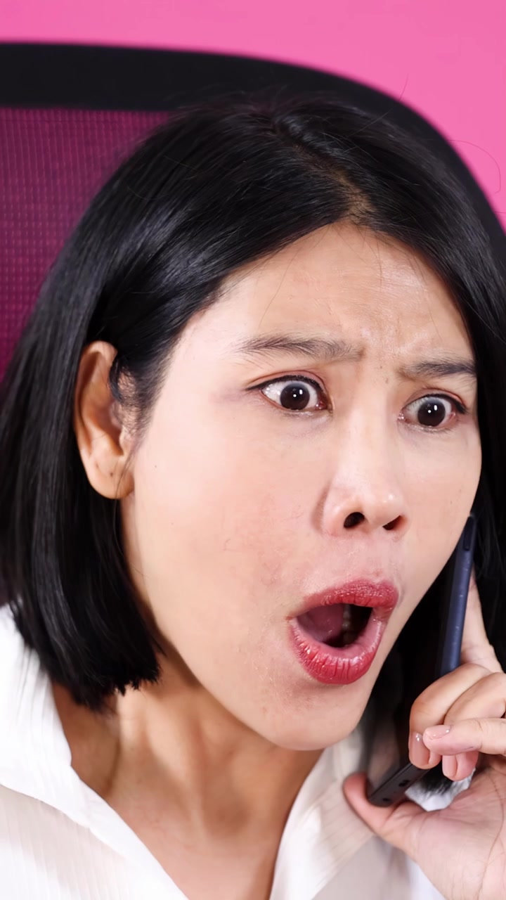 분홍색 사무실 배경에서 전화 통화 중 놀라움과 분노를 드러내는 흰색 셔츠를 입은 아시아 여성의 클로즈업.