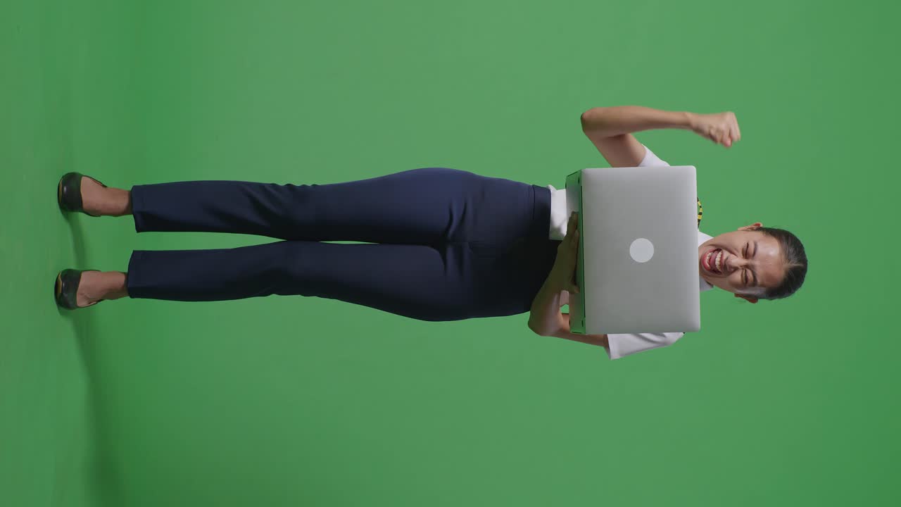 아시안 여성 조종사가 노트북을 바라보며 소리를 지르며 녹색 스크린 배경 스튜디오에 서서 골을 축하하고 있습니다.