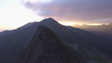 Jagged-mountain-peak-and-narrow-ridge-to-summit-with-dramatic-sky-and-golden-light-rays-streaking-across-the-sky-in-the-distance