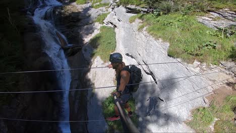 young and athletic man is walking over on a very small rope on via ferrata hiking route with a small and beautiful river waterfall running next to and under him. holding on carefully to not fall down.