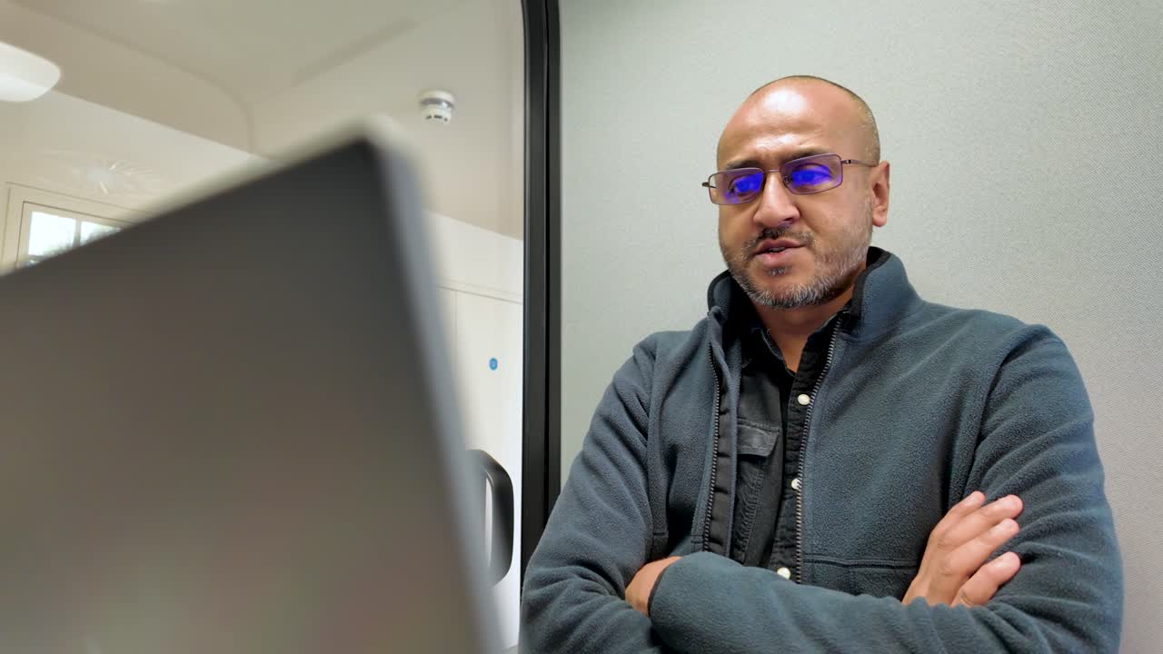 South asian office worker attending a webinar in office pod, with arms crossed, swiveling in chair