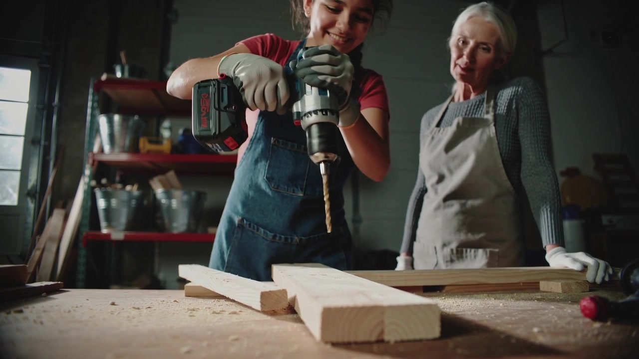 Mentor guiding red-shirted teen in overalls using cordless drill on plank at bench, making shavings