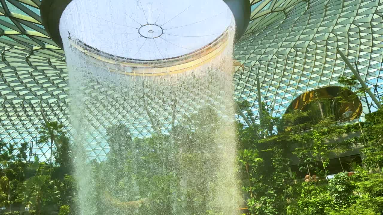 A dramatic indoor waterfall pours from a glass dome into a verdant garden atrium, with natural daylight illuminating tropical plants and architectural details. Subtle camera movement reveals changing perspectives
