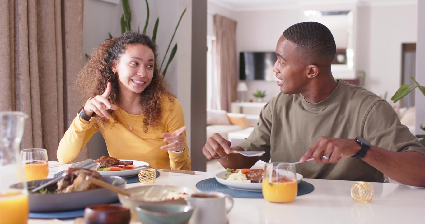 Couple sitting at home dining table, conversing, woman speaking, man cutting on plate with fork