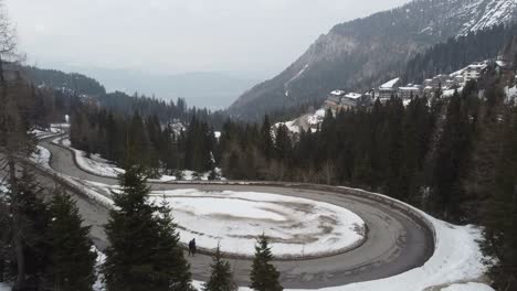 Aufschlussreiche-Aufnahme-Einer-Verschneiten-Serpentinenstraße-In-Den-Bergen-Und-Eines-Ski-Hotelresorts-Mit-Zwei-Anonymen-Reisenden,-Die-Spazieren-Gehen