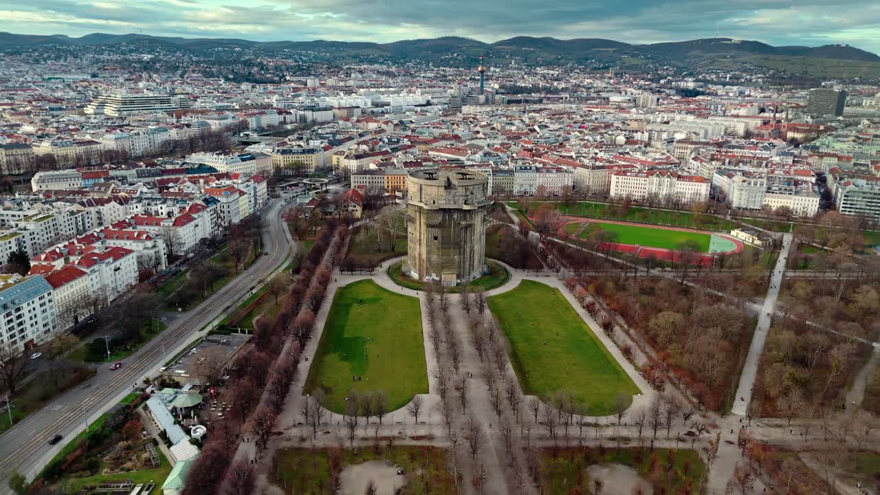 Laaja ilmakuva drone-otoksena Wienin Augarten-tornista talvisessa auringonlaskussa, jossa näkyy pilvenpiirtäjä, ympäröivät puistoalueet, paljaat puut, kaupungin rakennukset ja lämmin auringonvalo.