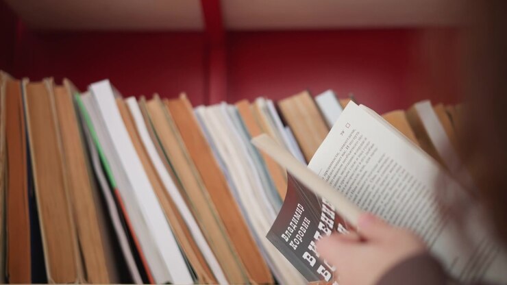 Una persona sfoglia vari titoli di libri prendendo diversi volumi per esaminarli. L'inquadratura si concentra sull'interazione con i materiali stampati davanti a uno scaffale con sfondo rosso