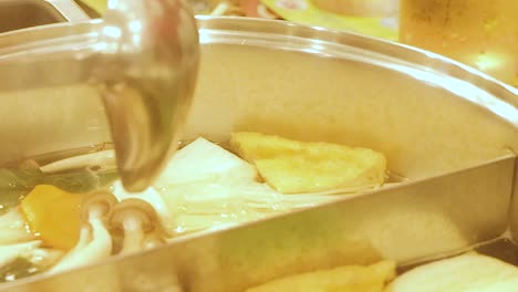 close-up of a ladle stirring a hotpot with tofu, mushrooms, and vegetables in a simmering broth.