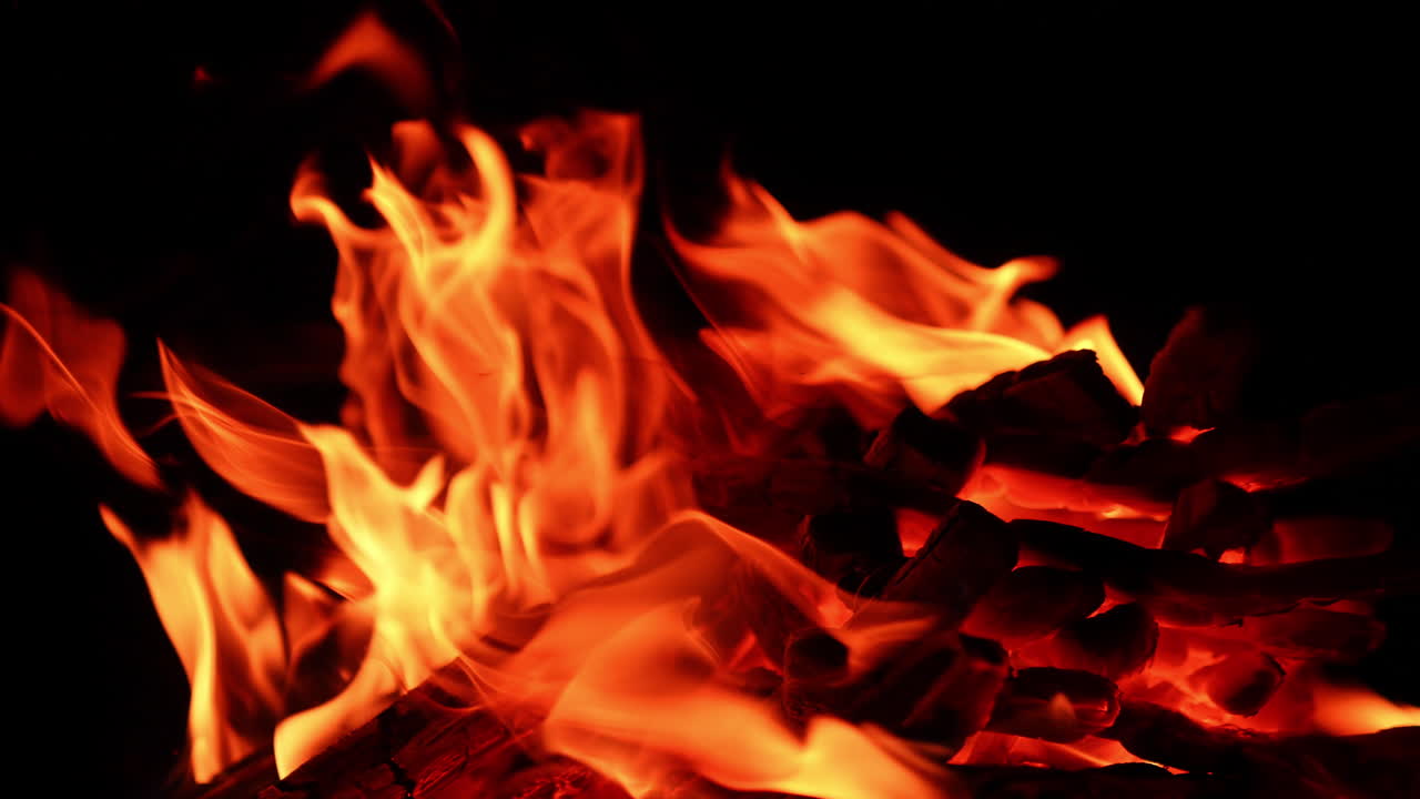 Tronchi di legno accatastati bruciano nel falò. Fiamme calde si muovono nell'oscurità, in primo piano.